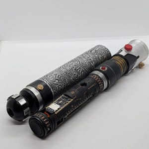 May include: A cylindrical lightsaber hilt with a black and white swirling patterned grip, alongside a second hilt featuring a black chassis with exposed copper-colored circuitry, a small red button, and a silver emitter with a red activation switch.