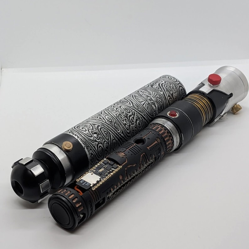 May include: A cylindrical lightsaber hilt with a black and white swirling patterned grip, alongside a second hilt featuring a black chassis with exposed copper-colored circuitry, a small red button, and a silver emitter with a red activation switch.