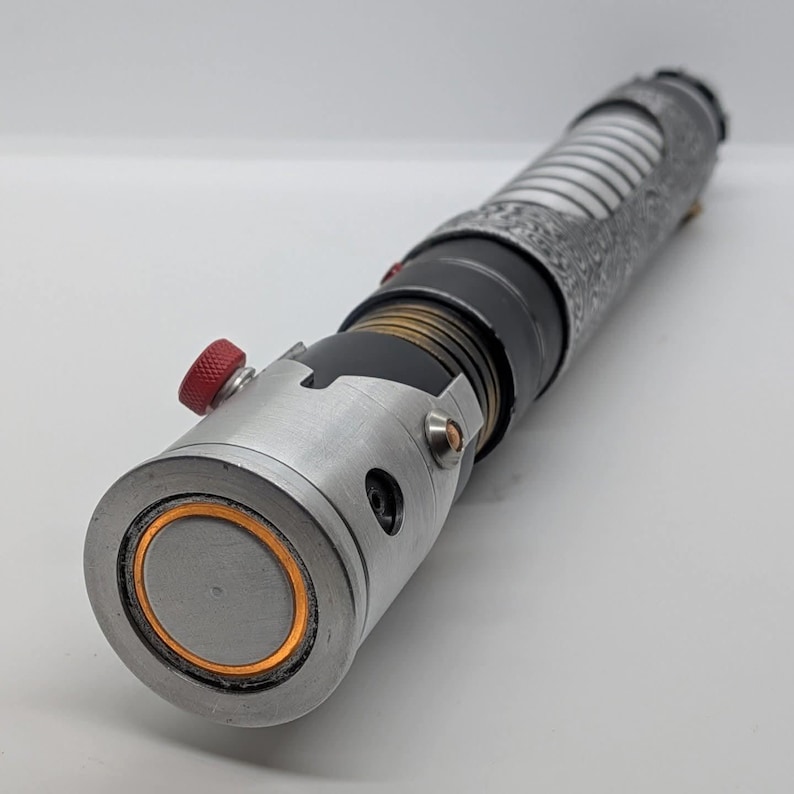 May include: A cylindrical metal lightsaber hilt featuring a brushed silver emitter with an orange inner ring, a black midsection with brass-colored ribbed detailing, and a textured silver grip. A small red knurled button is visible on the side.
