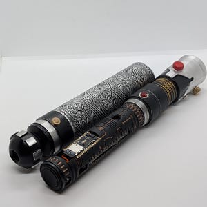 May include: A disassembled lightsaber hilt featuring a black and silver patterned outer sleeve with a swirling design. The inner core is black with intricate copper-colored mechanical detailing, exposed circuitry, a small red button, and a silver emitter.