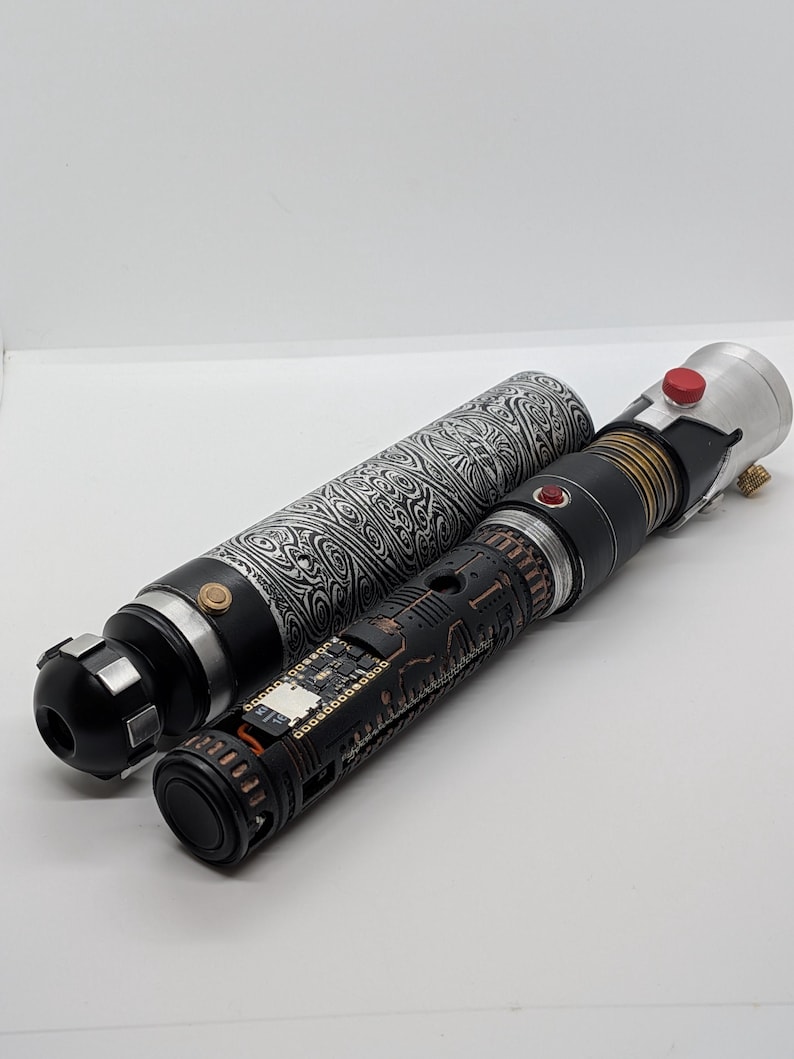 May include: A disassembled lightsaber hilt featuring a black and silver patterned outer sleeve with a swirling design. The inner core is black with intricate copper-colored mechanical detailing, exposed circuitry, a small red button, and a silver emitter.