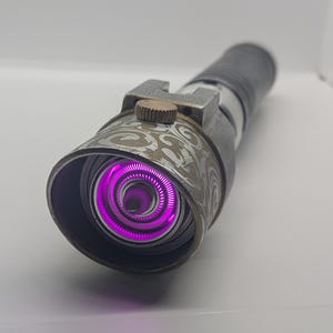 May include: A cylindrical metal prop with a weathered grey finish and intricate swirling patterns on the emitter shroud. The device features a bright, glowing purple circular light at the center and a small, textured bronze-colored adjustment knob.