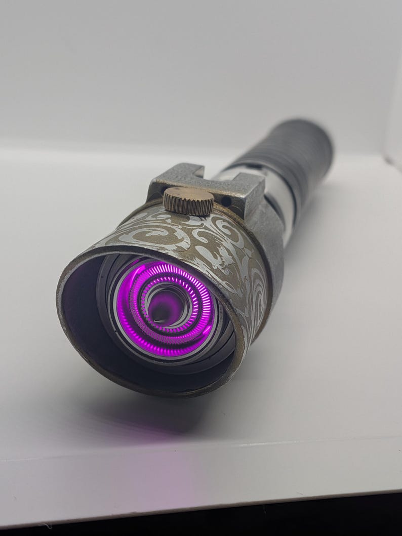 May include: A cylindrical metal prop with a weathered grey finish and intricate swirling patterns on the emitter shroud. The device features a bright, glowing purple circular light at the center and a small, textured bronze-colored adjustment knob.