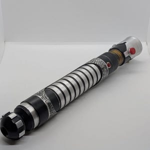 May include: A cylindrical lightsaber hilt featuring a matte black finish with silver ribbed sections and intricate etched patterns. The handle includes a gold-toned neck, a prominent red activation button, and a small red accent stud on the side.