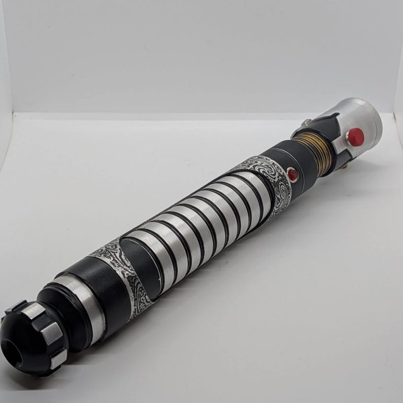 May include: A cylindrical lightsaber hilt featuring a matte black finish with silver ribbed sections and intricate etched patterns. The handle includes a gold-toned neck, a prominent red activation button, and a small red accent stud on the side.