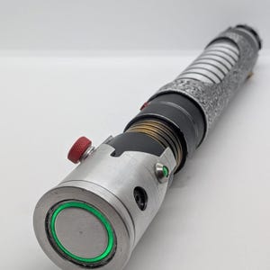 May include: A cylindrical metal prop with a silver emitter featuring a glowing green ring, a red knurled activation button, and a small green accent light. The handle includes a black section with bronze-colored rings and a patterned silver grip.