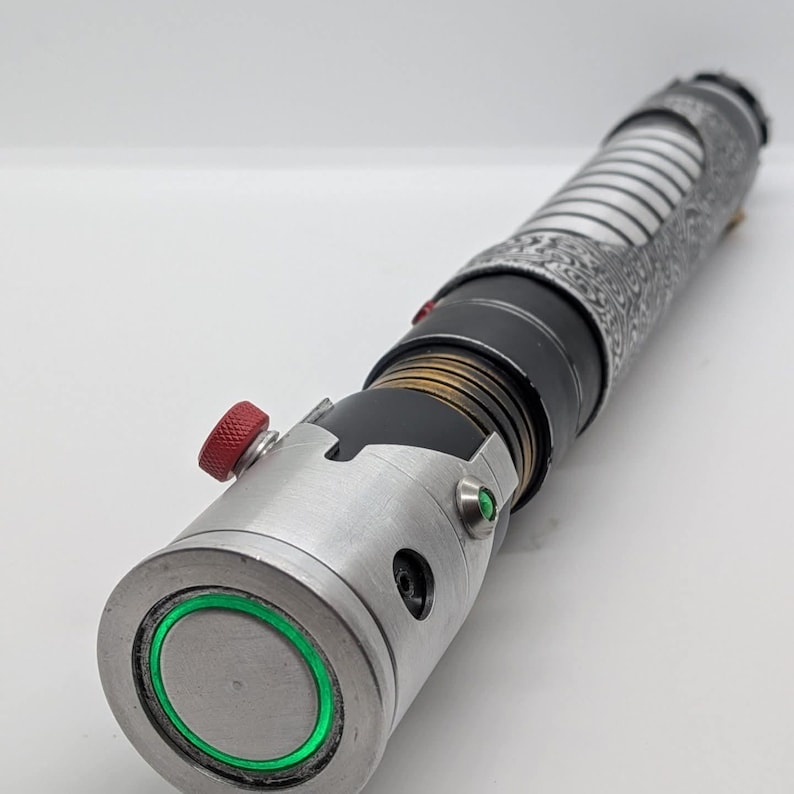 May include: A cylindrical metal prop with a silver emitter featuring a glowing green ring, a red knurled activation button, and a small green accent light. The handle includes a black section with bronze-colored rings and a patterned silver grip.