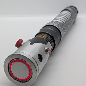 May include: A cylindrical metal prop with a silver finish, featuring a textured grip section, a black midsection with brass-colored rings, and a silver emitter end with a bright red circular light accent and a small red knurled adjustment knob.