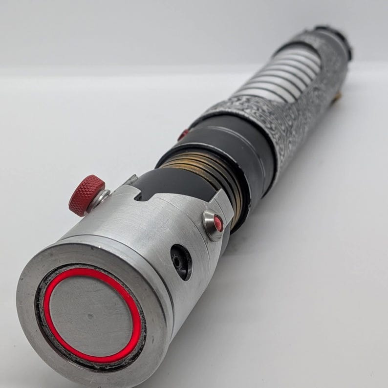May include: A cylindrical metal prop with a silver finish, featuring a textured grip section, a black midsection with brass-colored rings, and a silver emitter end with a bright red circular light accent and a small red knurled adjustment knob.