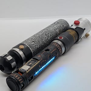 May include: A cylindrical lightsaber hilt with a black and white swirling Damascus steel pattern, paired with a second hilt featuring a matte black finish, copper-colored accents, a small red button, and a glowing blue light strip along the side.