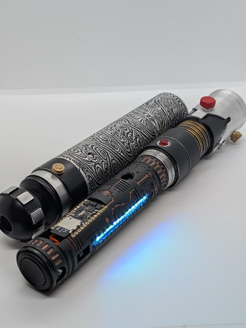 May include: A cylindrical lightsaber hilt with a black and white swirling Damascus steel pattern, paired with a second hilt featuring a matte black finish, copper-colored accents, a small red button, and a glowing blue light strip along the side.
