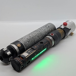 May include: A cylindrical prop with a black and white swirling pattern next to a black metal handle featuring exposed electronic components, a glowing green light strip, a small red button, and a silver top section with a red circular button.