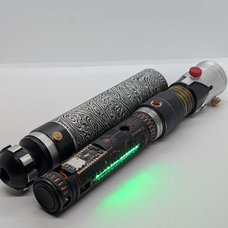 May include: A cylindrical prop with a black and white swirling pattern next to a black metal handle featuring exposed electronic components, a glowing green light strip, a small red button, and a silver top section with a red circular button.