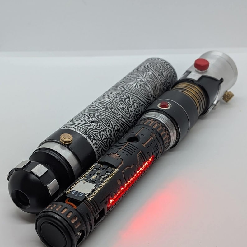 May include: A cylindrical metal prop with a black and silver patterned finish alongside a second cylindrical prop featuring a black textured exterior, exposed electronic circuitry, a small red light, and a prominent red button on the silver top.