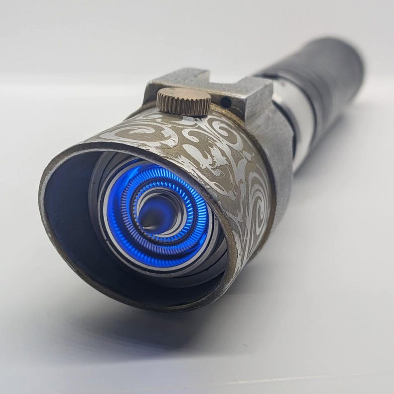 May include: A cylindrical metal prop with a weathered finish and an intricate etched scrollwork pattern on the emitter shroud. A small bronze-colored dial sits atop the shroud, while the interior features a glowing, vibrant blue circular light.