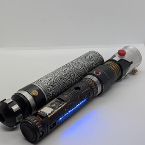 May include: A cylindrical lightsaber hilt with a black and white damascus pattern sleeve, featuring a matte black inner chassis with exposed electronic components, a glowing blue LED strip, a red activation button, and metallic silver accents.