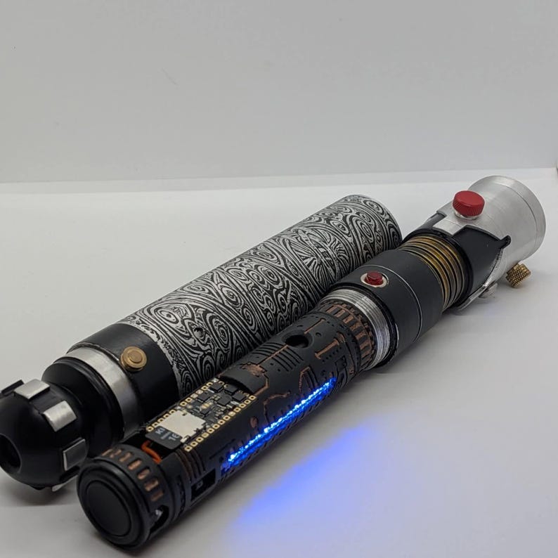 May include: A cylindrical lightsaber hilt with a black and white damascus pattern sleeve, featuring a matte black inner chassis with exposed electronic components, a glowing blue LED strip, a red activation button, and metallic silver accents.