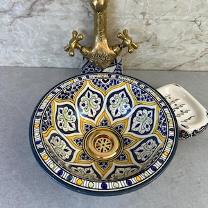 May include: A round ceramic vessel sink featuring a vibrant hand-painted floral pattern in shades of deep blue, yellow, and white. The sink includes a polished brass drain assembly and is paired with an ornate, antique-style brass faucet.