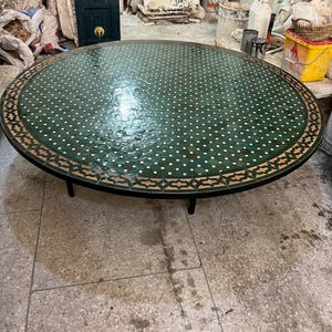 May include: A large round table featuring a green mosaic tile top with a repeating white geometric pattern and a decorative border of tan and dark green tiles. The table is supported by a black metal frame with thin, cylindrical legs.