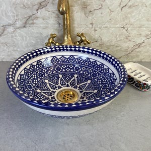 Peut inclure: Une vasque ronde en céramique ornée d'un motif géométrique et floral bleu et blanc avec un bord à carreaux. La vasque comprend une bonde en métal doré et est accompagnée d'un robinet doré assorti et d'un petit porte-savon rectangulaire.