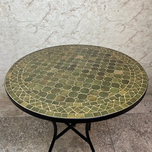 May include: A round table featuring a mosaic top composed of various shades of green tiles in octagonal and diamond shapes. The tabletop is framed by a thin black metal edge and is supported by a black metal base with four curved legs.