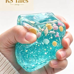 May include: A squishy, translucent turquoise cube filled with small, colourful seashell figurines and tiny white star-shaped glitter. The material is flexible and pliable, allowing it to be compressed while maintaining its overall square shape.