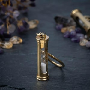May include: A brass hourglass keychain featuring a cylindrical metal frame with a clear glass center containing white sand. The top of the brass casing has a small loop attached to a circular metal ring, allowing it to function as a functional keychain.