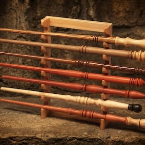 May include: A collection of seven wooden wands with varied grain patterns and shades of brown, ranging from light tan to deep reddish-brown, displayed horizontally on a light-colored wooden rack with notched supports for each individual wand.