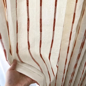 May include: A cream-coloured curtain featuring vertical stripes in a deep rust-orange hue. The fabric displays a subtle, tone-on-tone woven pattern throughout, with the rust-coloured stripes accented by small, horizontal, dashed-line details.