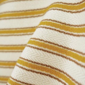 May include: A woven textile featuring a repeating pattern of horizontal mustard yellow and thin brown stripes set against an off-white background. The fabric displays a distinct herringbone weave texture throughout the entire surface area.