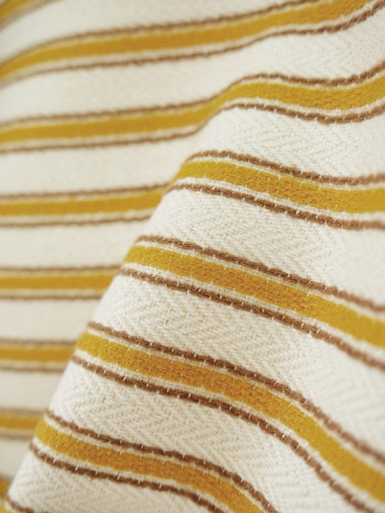 May include: A woven textile featuring a repeating pattern of horizontal mustard yellow and thin brown stripes set against an off-white background. The fabric displays a distinct herringbone weave texture throughout the entire surface area.