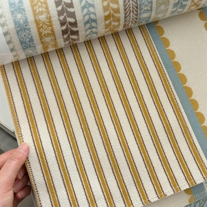 May include: A fabric sample book featuring a page with vertical mustard yellow and dark brown stripes on an off-white background. The top section displays a horizontal band of patterned fabric with blue, yellow, and brown floral and leaf motifs.