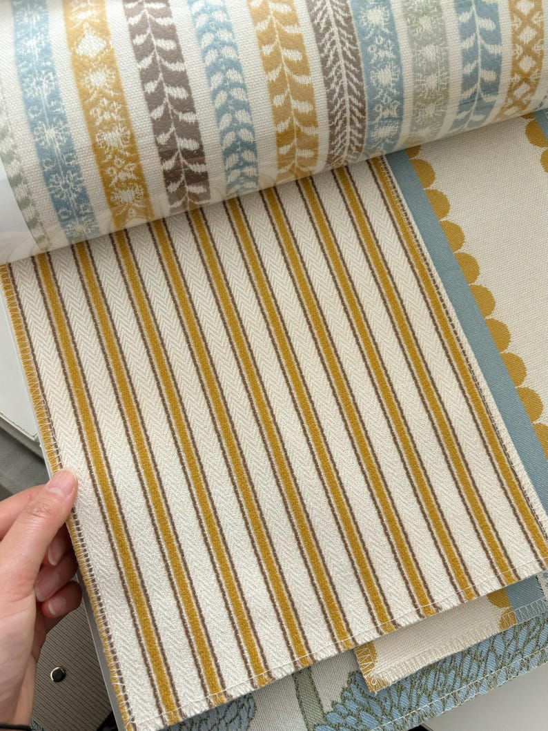May include: A fabric sample book featuring a page with vertical mustard yellow and dark brown stripes on an off-white background. The top section displays a horizontal band of patterned fabric with blue, yellow, and brown floral and leaf motifs.