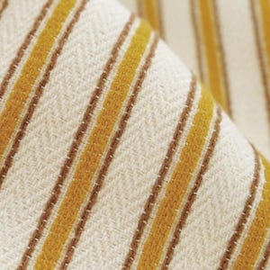 May include: A woven fabric featuring a repeating pattern of vertical mustard yellow stripes separated by thin brown lines against an off-white background. The textile displays a distinct herringbone weave texture throughout the entire surface.