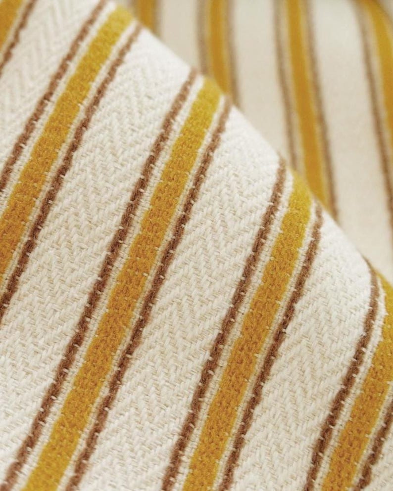 May include: A woven fabric featuring a repeating pattern of vertical mustard yellow stripes separated by thin brown lines against an off-white background. The textile displays a distinct herringbone weave texture throughout the entire surface.