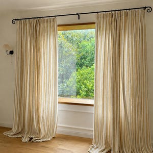 May include: Floor-length curtains featuring a vertical striped pattern in shades of mustard yellow and off-white. The fabric hangs from a black metal curtain rod with decorative scroll ends, creating soft folds that extend to the floor.