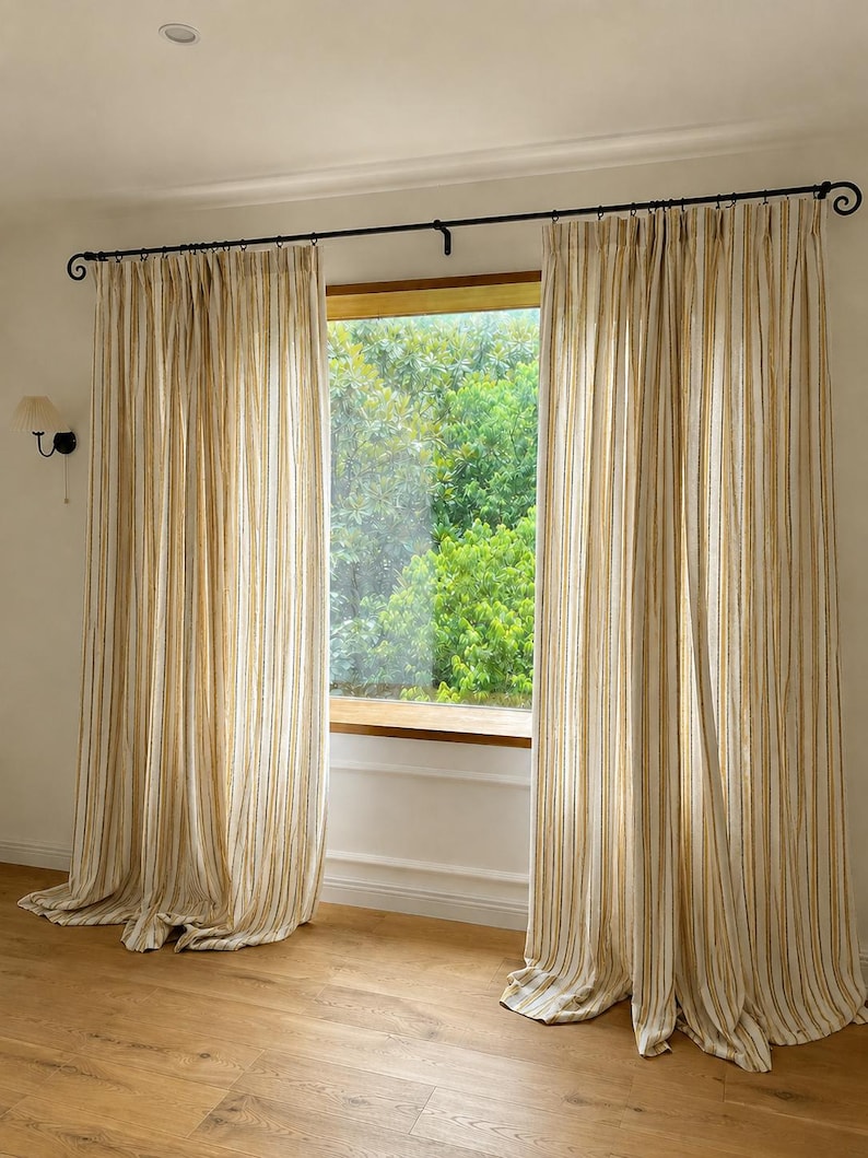 May include: Floor-length curtains featuring a vertical striped pattern in shades of mustard yellow and off-white. The fabric hangs from a black metal curtain rod with decorative scroll ends, creating soft folds that extend to the floor.