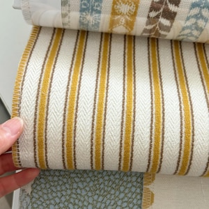 May include: A fabric sample book featuring a prominent swatch with vertical mustard yellow and brown stripes on a cream background, layered over other swatches displaying blue and yellow patterned designs and a textured light blue fabric.