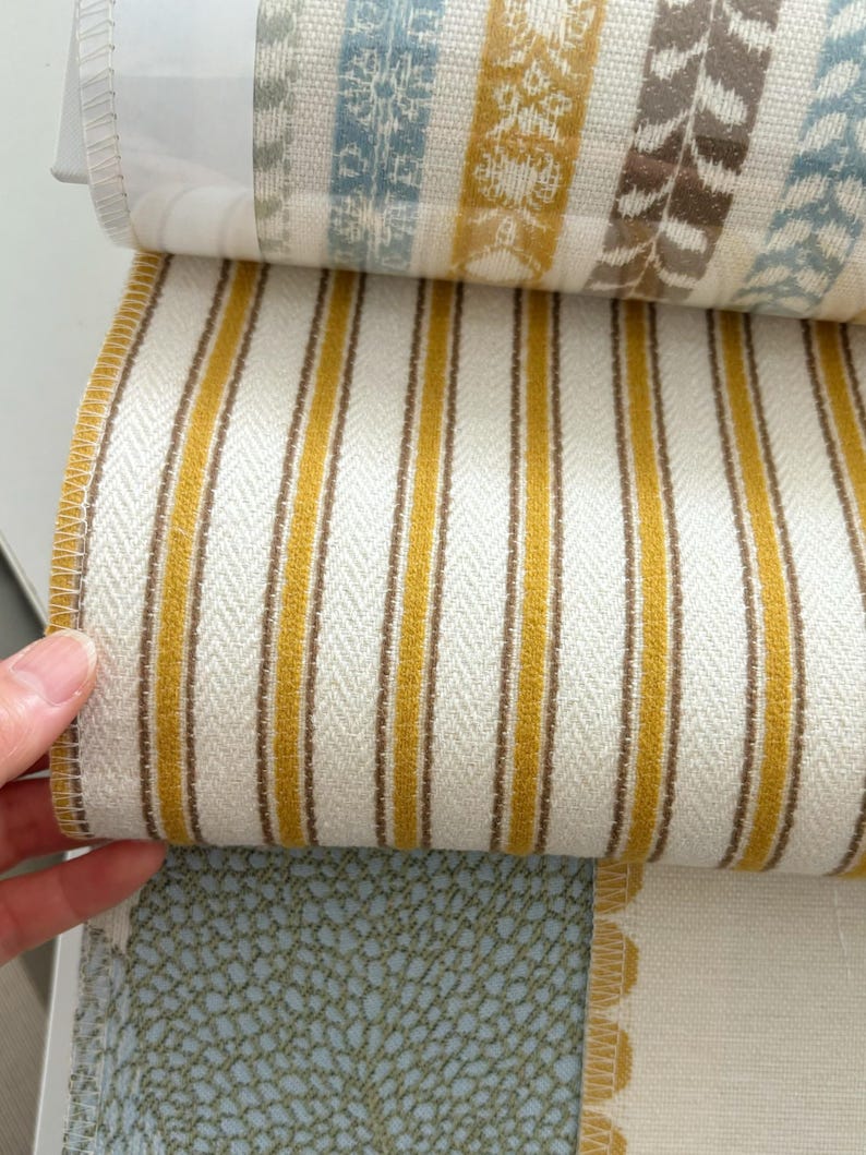 May include: A fabric sample book featuring a prominent swatch with vertical mustard yellow and brown stripes on a cream background, layered over other swatches displaying blue and yellow patterned designs and a textured light blue fabric.