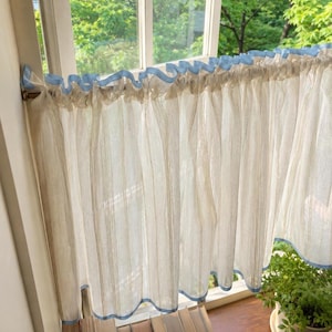 May include: A sheer, off-white cafe curtain featuring vertical textured stripes and a light blue trim along the top ruffle and bottom hem. The curtain is hung on a thin rod, displaying a gathered, pleated design that allows light to pass through.