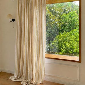 May include: A floor-length curtain featuring vertical stripes in shades of mustard yellow, white, and light gray. The curtain is suspended from a black metal rod with decorative scroll ends and hangs in soft, vertical folds covering a window.