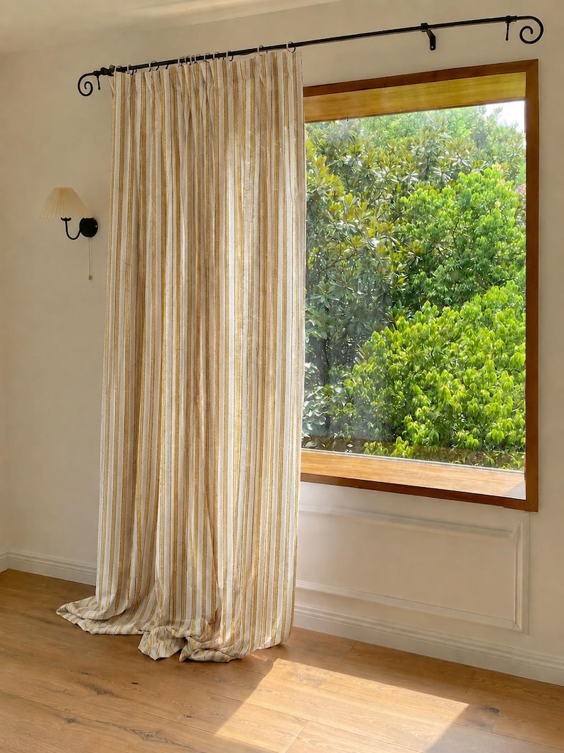May include: A floor-length curtain featuring vertical stripes in shades of mustard yellow, white, and light gray. The curtain is suspended from a black metal rod with decorative scroll ends and hangs in soft, vertical folds covering a window.