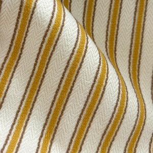 May include: A woven fabric featuring a repeating pattern of vertical mustard yellow and thin brown stripes set against an off-white background. The textile displays a distinct herringbone weave texture throughout, with soft folds creating depth and shadows.