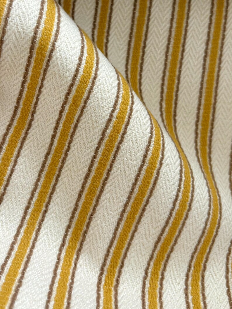 May include: A woven fabric featuring a repeating pattern of vertical mustard yellow and thin brown stripes set against an off-white background. The textile displays a distinct herringbone weave texture throughout, with soft folds creating depth and shadows.