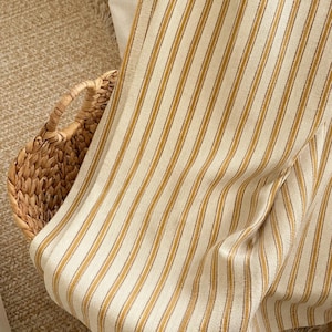 May include: A woven fabric textile featuring a repeating pattern of vertical mustard yellow and thin grey stripes against an off-white background. The fabric is draped over a woven wicker basket with visible handles, showcasing the texture of the weave.
