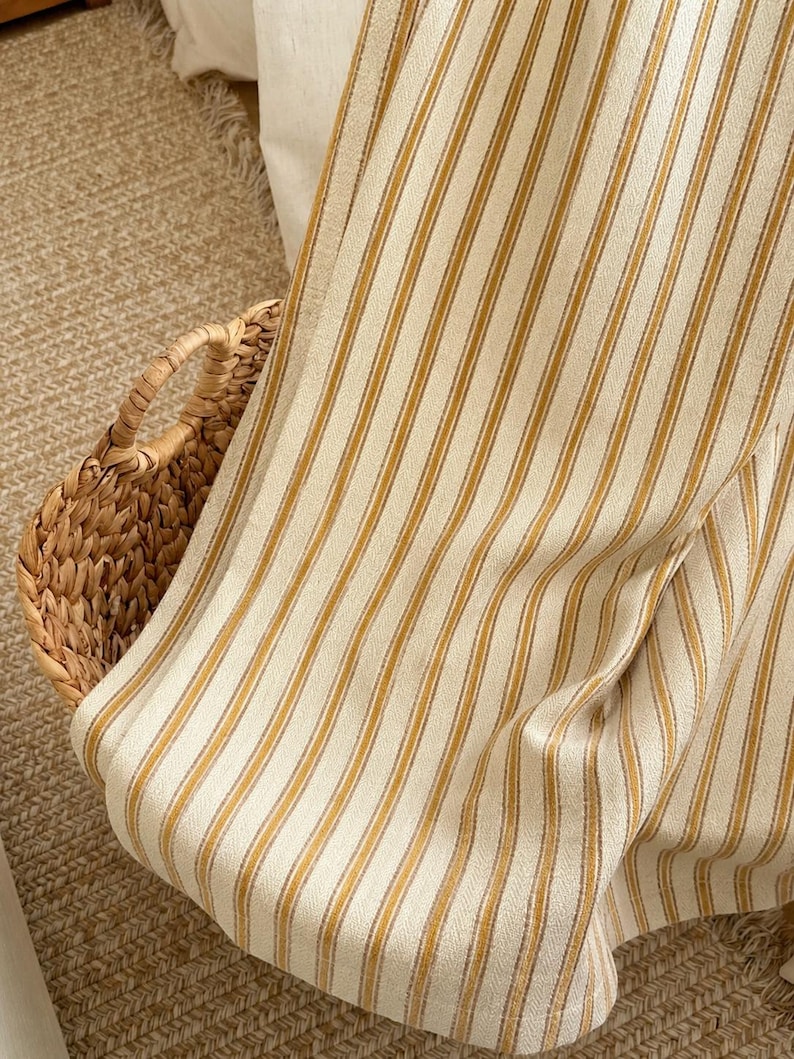 May include: A woven fabric textile featuring a repeating pattern of vertical mustard yellow and thin grey stripes against an off-white background. The fabric is draped over a woven wicker basket with visible handles, showcasing the texture of the weave.