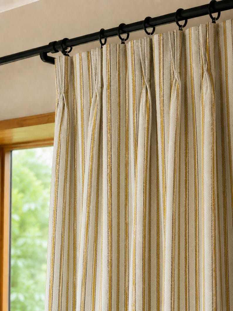 May include: A pinch-pleated curtain panel featuring a vertical striped pattern in shades of mustard yellow, white, and dark gray. The fabric hangs from a black metal curtain rod with matching black circular rings, creating uniform folds.