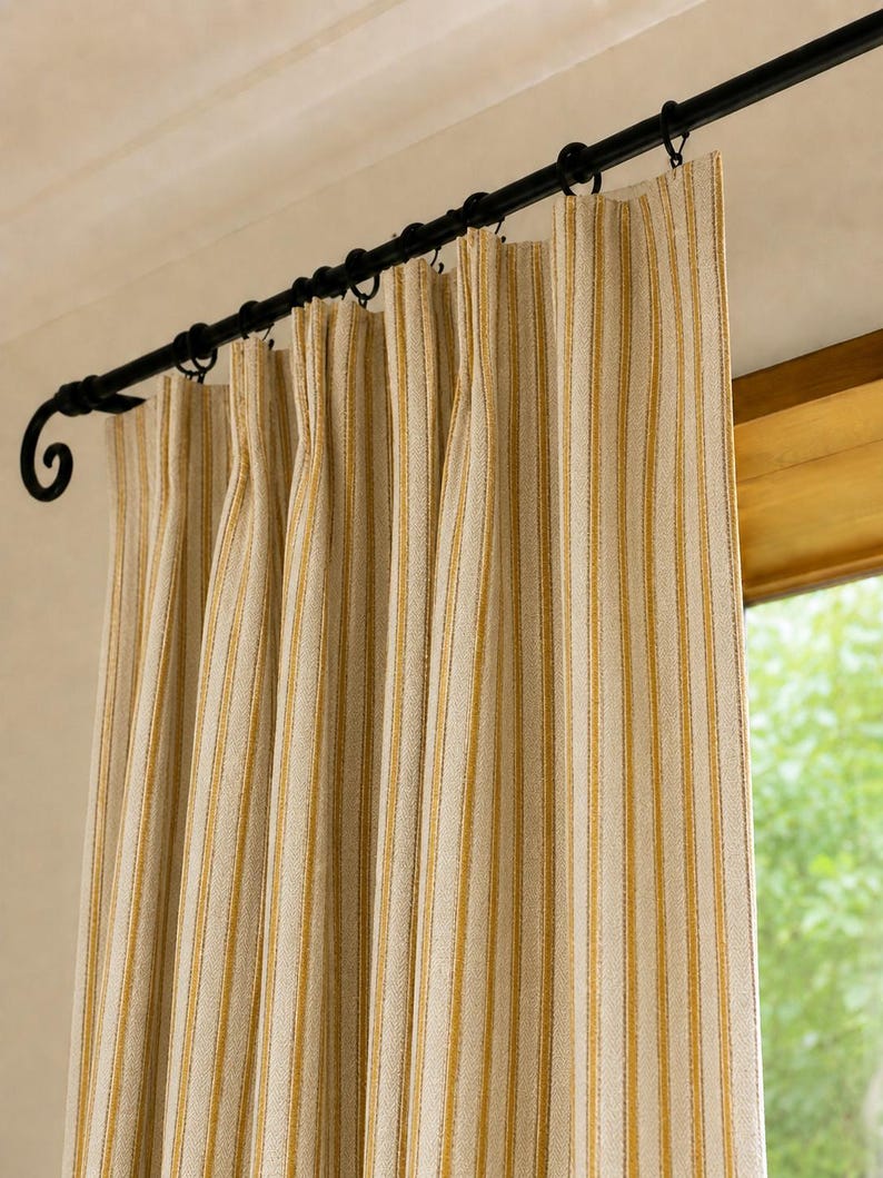 May include: A pleated curtain panel featuring a vertical striped pattern in mustard yellow and off-white. The fabric hangs from a black metal curtain rod with decorative scroll ends, secured by black circular rings that hold the fabric in place.