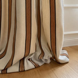 Peut inclure: Un rideau pleine longueur présentant un motif à rayures verticales dans des tons crème, beige et brun foncé. Le tissu présente une texture tissée visible et tombe en plis doux et fluides, s'accumulant légèrement au bas de la fenêtre.