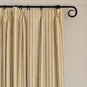 May include: A pair of off-white curtains featuring vertical mustard yellow stripes, suspended from a black metal rod with decorative scroll ends. The fabric is gathered into pleats at the top, creating a series of uniform vertical folds.